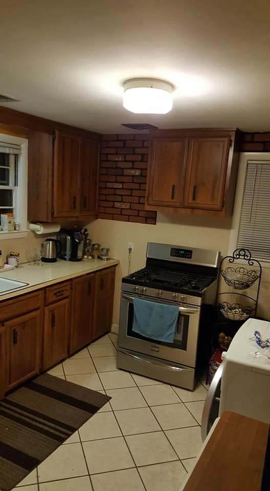 Kitchen with wooden cabinets, stainless steel stove, countertop with coffee maker and other appliances, brick wall, and window with blinds.