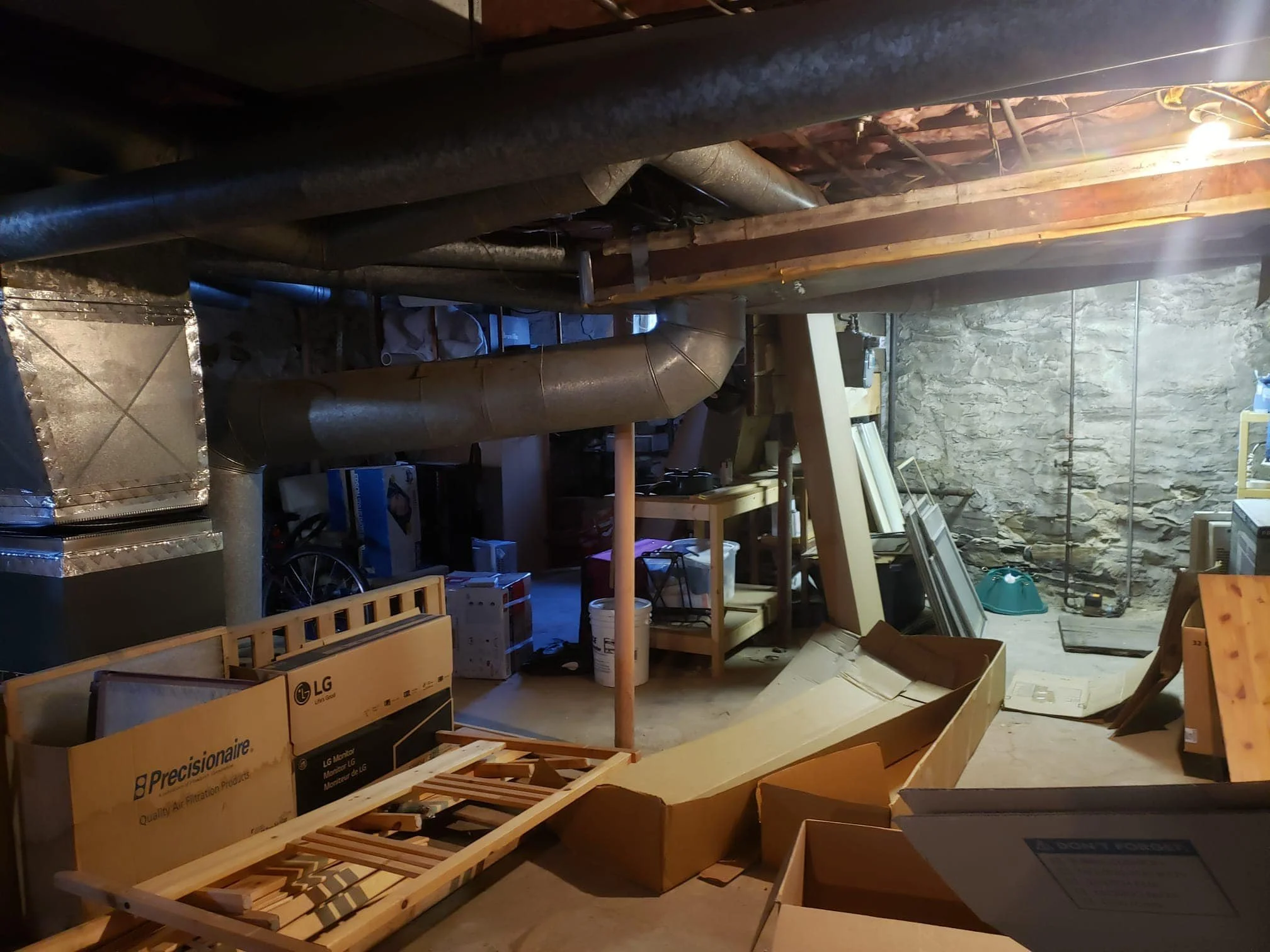 A cluttered basement with exposed brick wall, ventilation pipes, workbenches, storage boxes, unassembled furniture, and various tools and supplies scattered around.