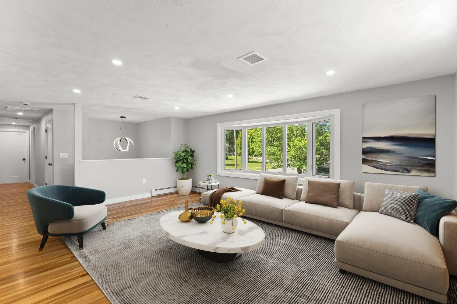 Bright living room with large window overlooking green trees, white sectional sofa with beige and brown pillows, teal armchair, round marble coffee table with yellow and black vases and flowers, gray area rug, potted plant, and abstract ocean-themed 