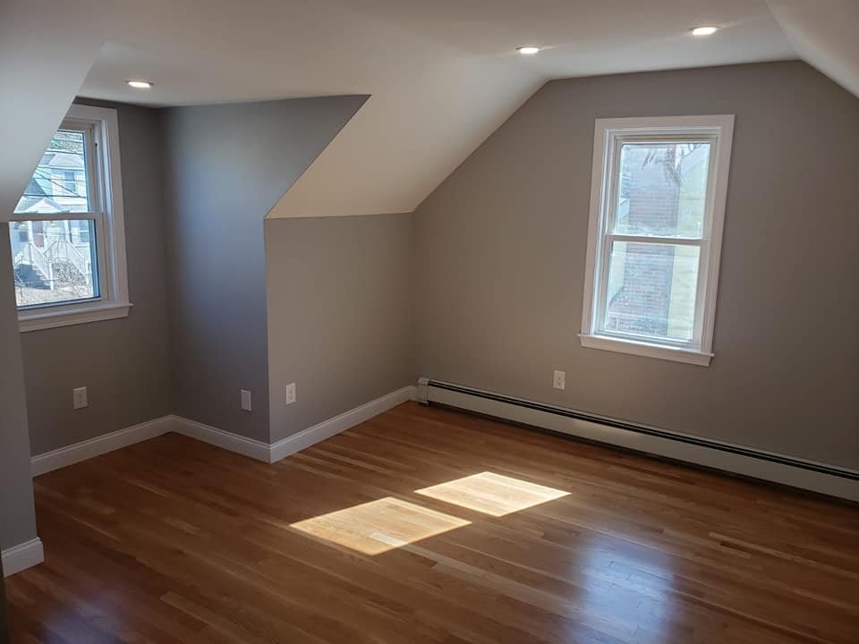 Empty room with hardwood floors, two windows letting in natural light, gray walls, and ceiling lights.