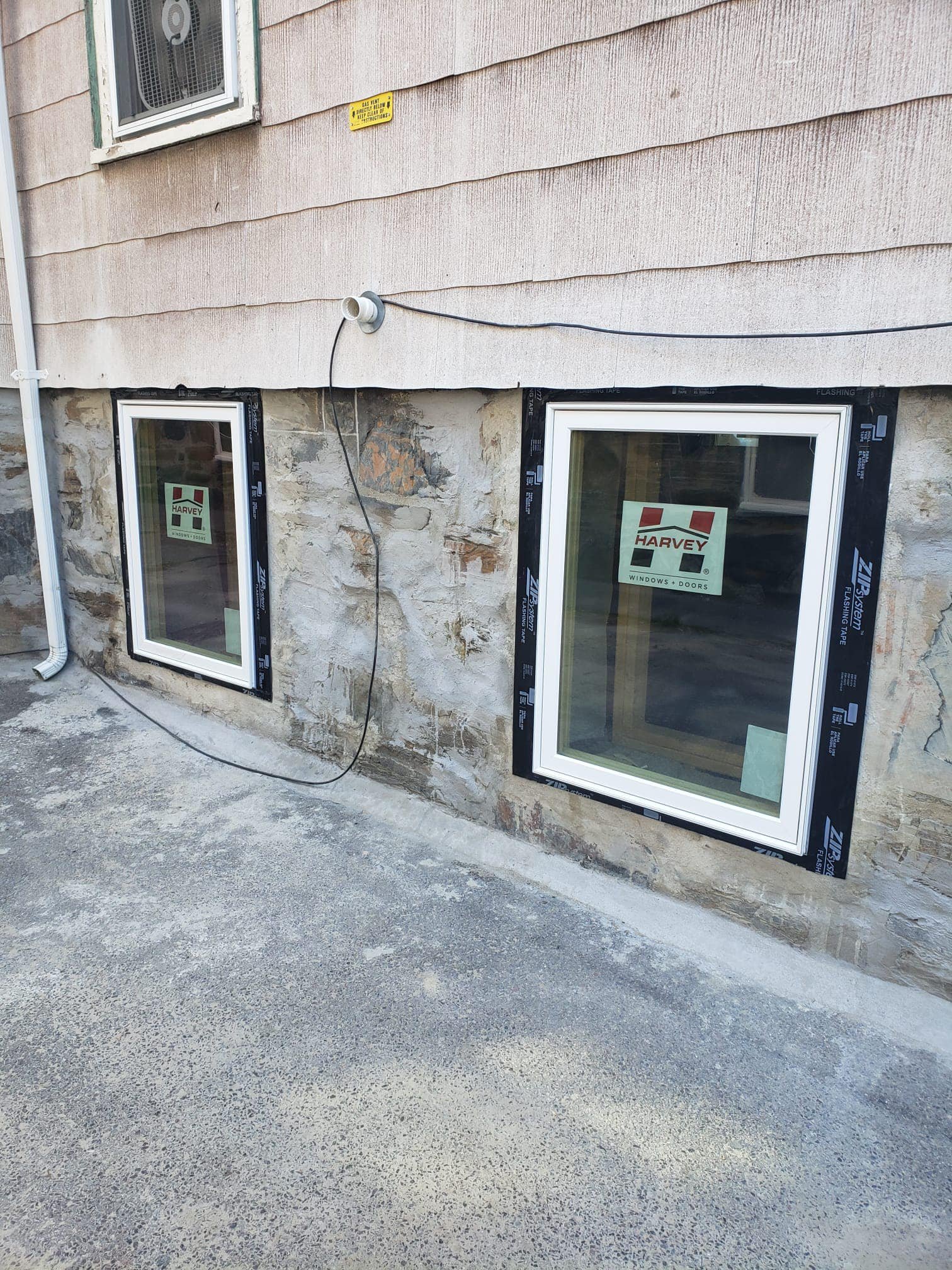 Construction site showing two new basement windows with Harvey sticker, stone foundation, and unfinished exterior wall with beige siding.