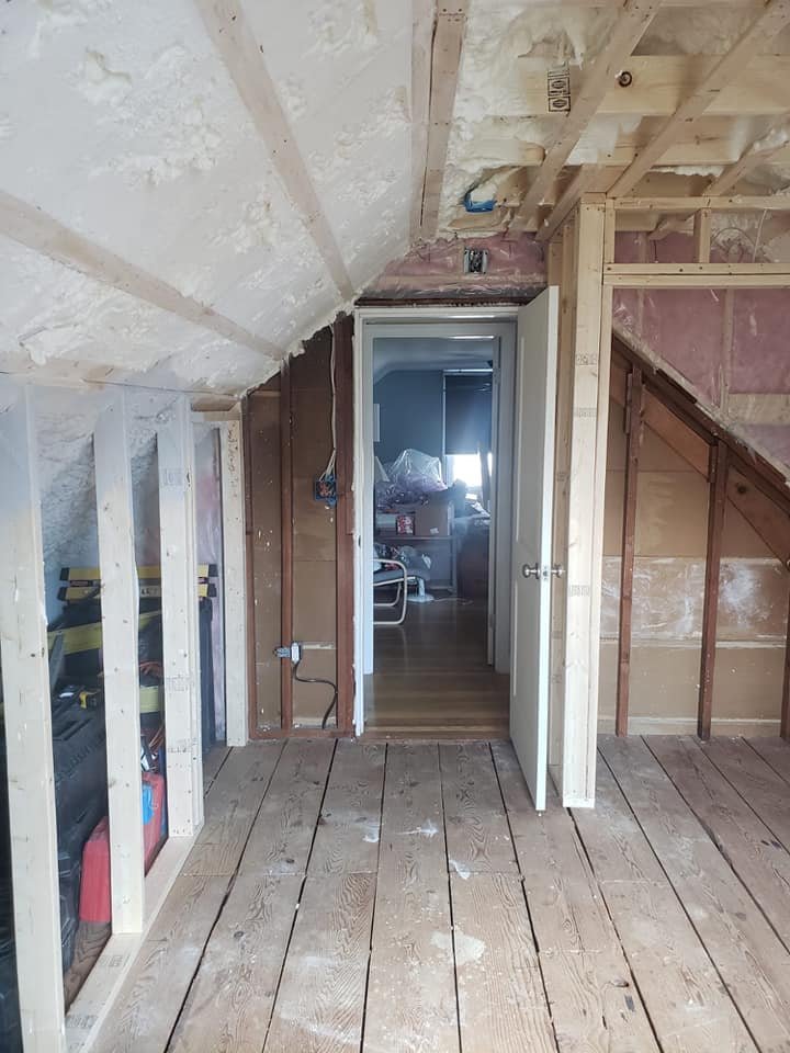 Interior of an attic or unfinished room with exposed wood framing and walls, a wooden floor with dust, and an open door leading to a room with a chair and a cluttered surface.