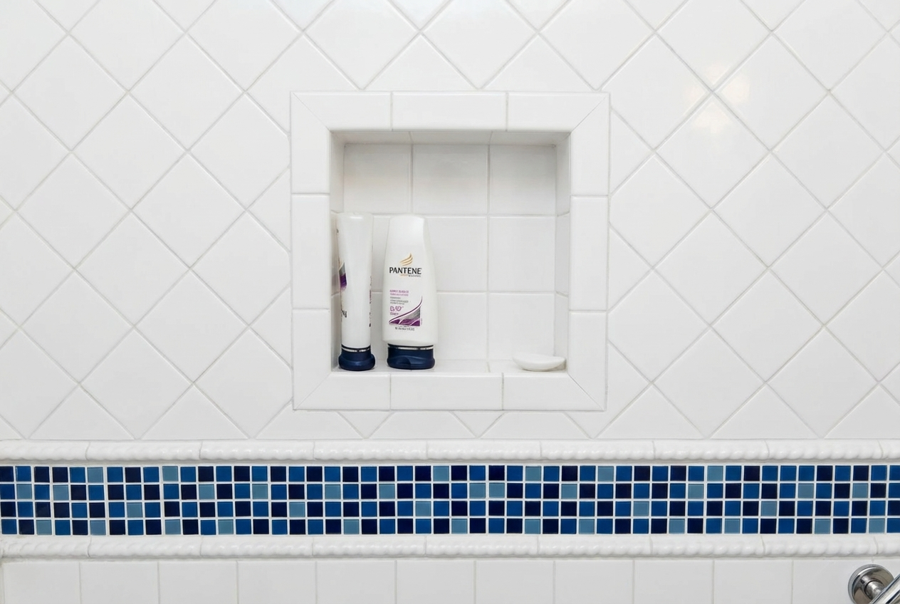 Close-up of a bathroom shower niche with two bottles of Pantene shampoo and a bar of soap, tiled with white ceramic and a horizontal band of blue mosaic tiles.