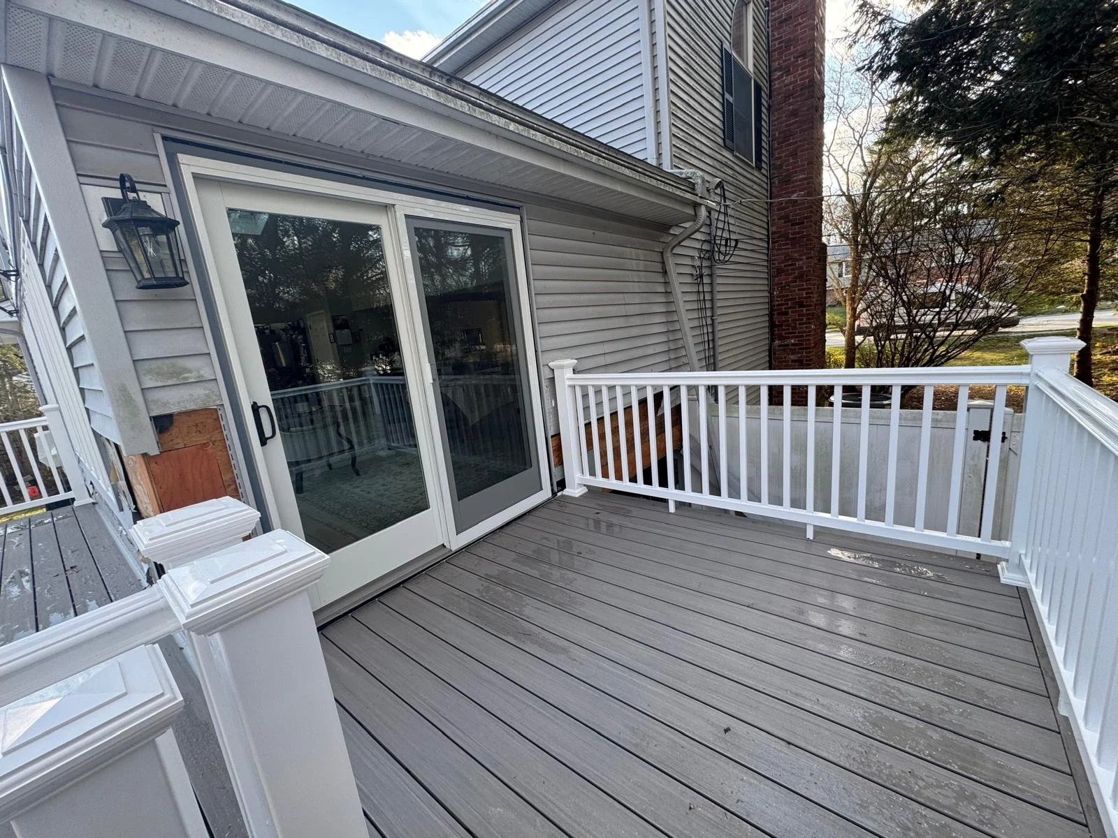 Empty outdoor wooden deck with white railing and sliding glass door leading into a house, with trees and neighboring house visible in the background.
