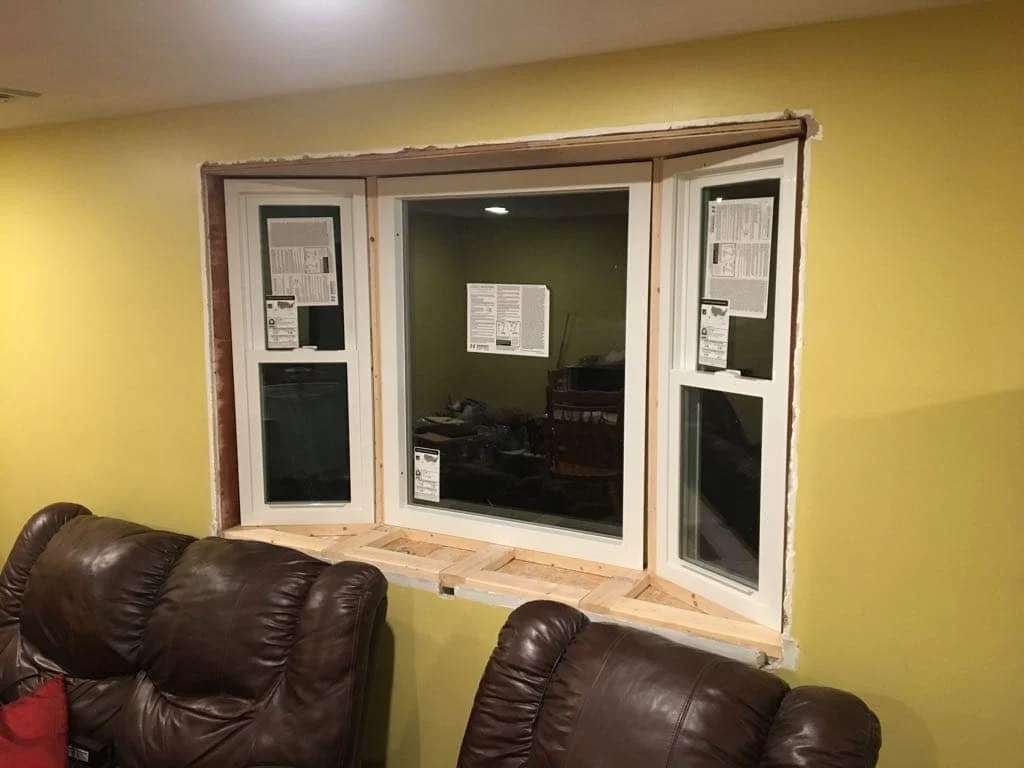 Interior of a room with yellow walls and a large three-pane window showing an unfinished window frame. Two brown leather couches are in the foreground.