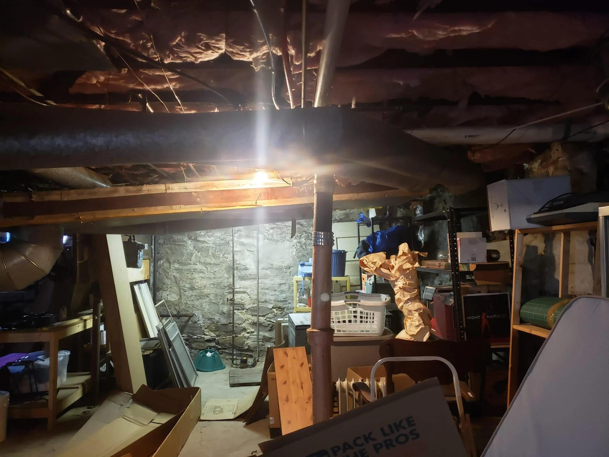 A cluttered basement with a stone wall in the background, packed with various shelves, boxes, and miscellaneous items, and a bright ceiling light illuminating the space.