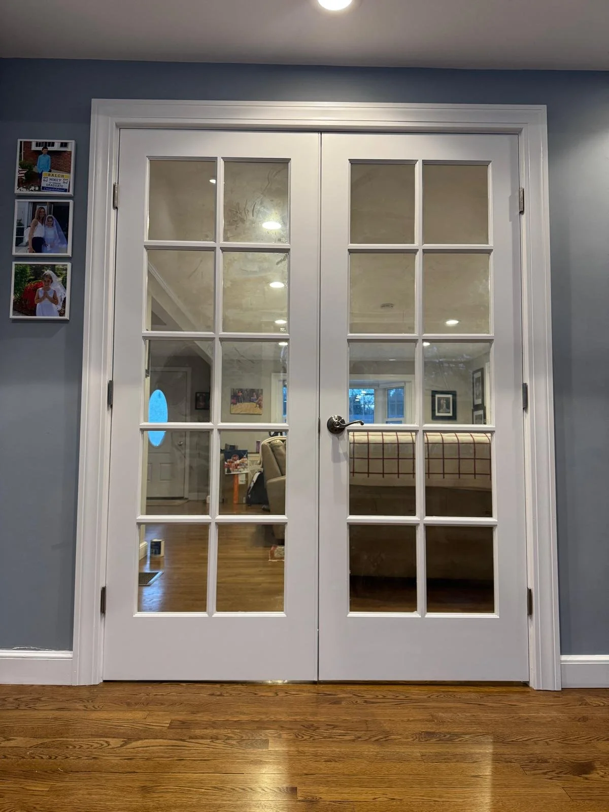 White double glass-paned French doors leading to a living room with wooden flooring, a beige sofa, and wall art, framed by gray walls and a hardwood floor.