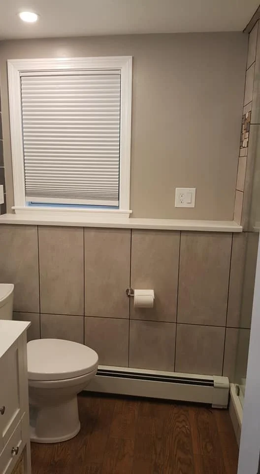Bathroom with beige tiled walls, a toilet, a window with blinds, a white baseboard heater, and a white outlet on the wall.