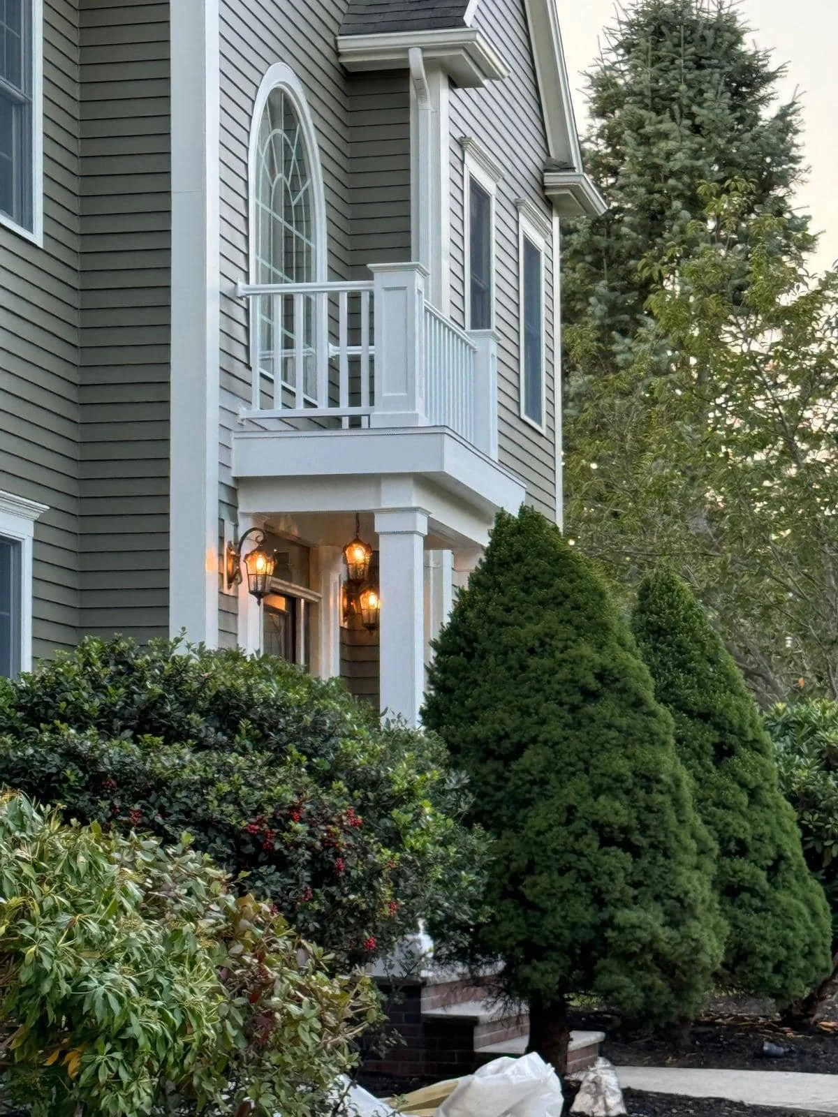 Front exterior of a house with beige siding, white trim, a small balcony with white railing, and multiple windows. There are lit outdoor lanterns by the entrance, and neatly trimmed bushes and trees around the house.