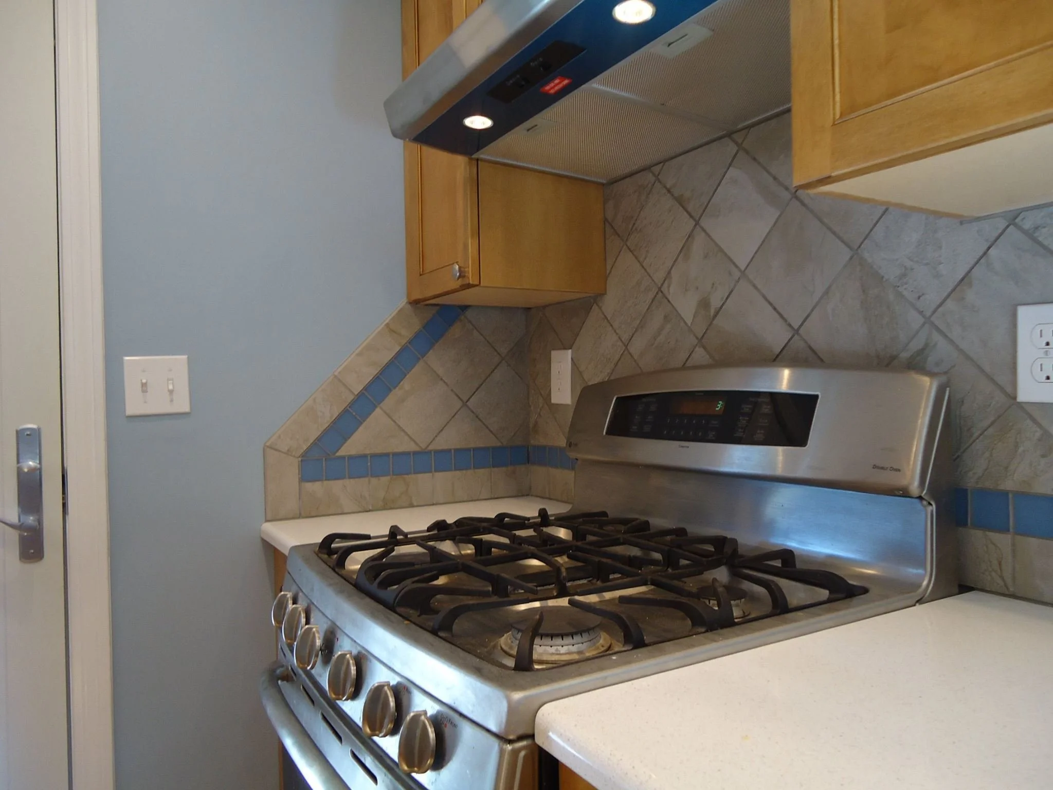 Stainless steel gas stove with four burners in a kitchen with beige tiled backsplash and light wooden cabinets.