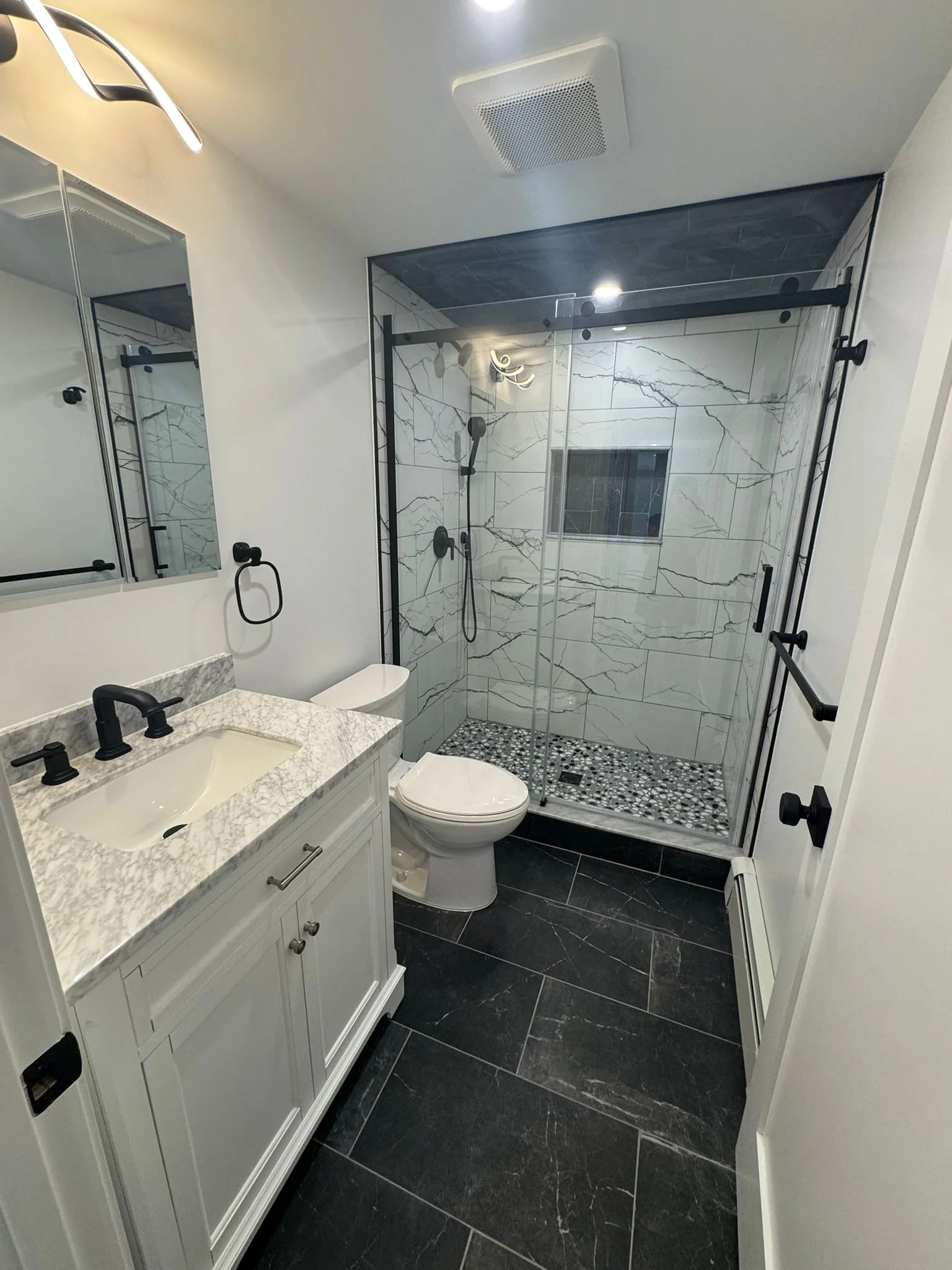 Modern bathroom with a white vanity with a marble countertop, a mirror, a toilet, and a walk-in shower with marble tiles and pebble flooring, black fixtures, and a glass sliding door.