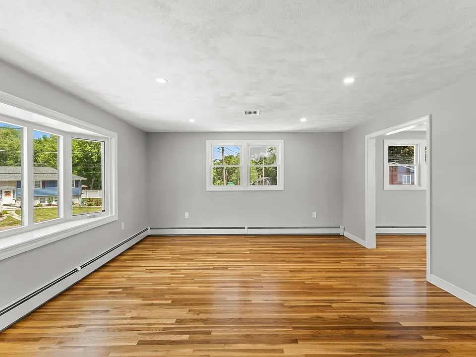 Empty living room with hardwood floors, white walls, ceiling lights, and multiple windows showing a neighborhood outside.