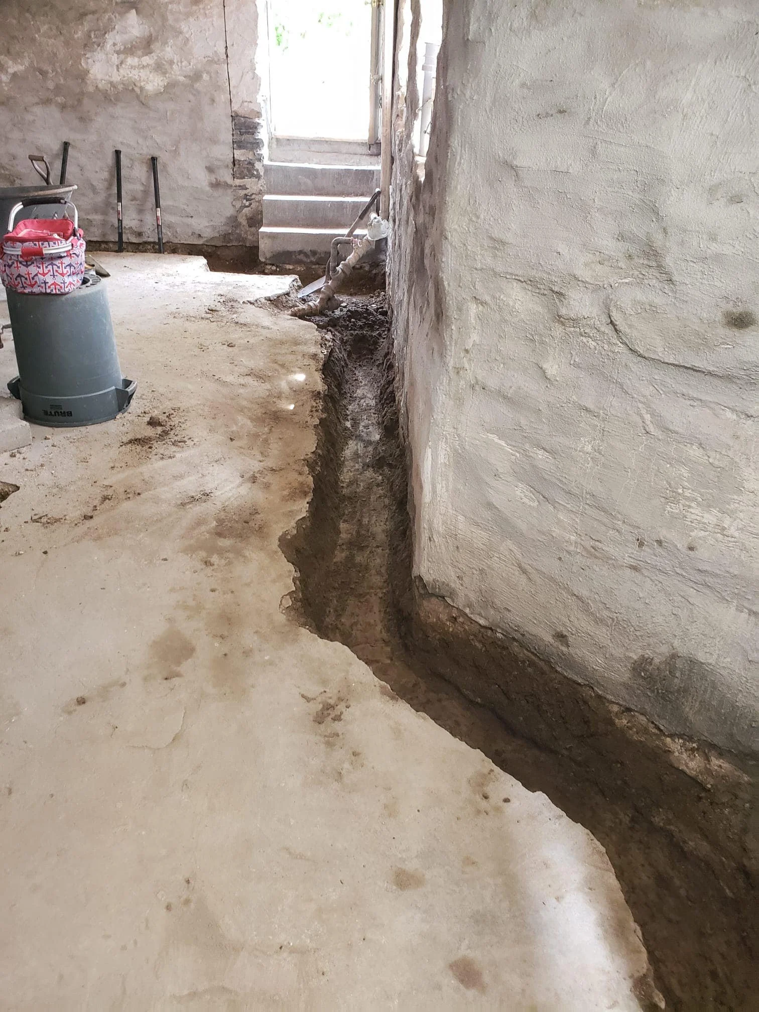 A construction site with a partially dug trench along the base of a wall, visible pipes near a doorway, and a bucket with cleaning supplies.