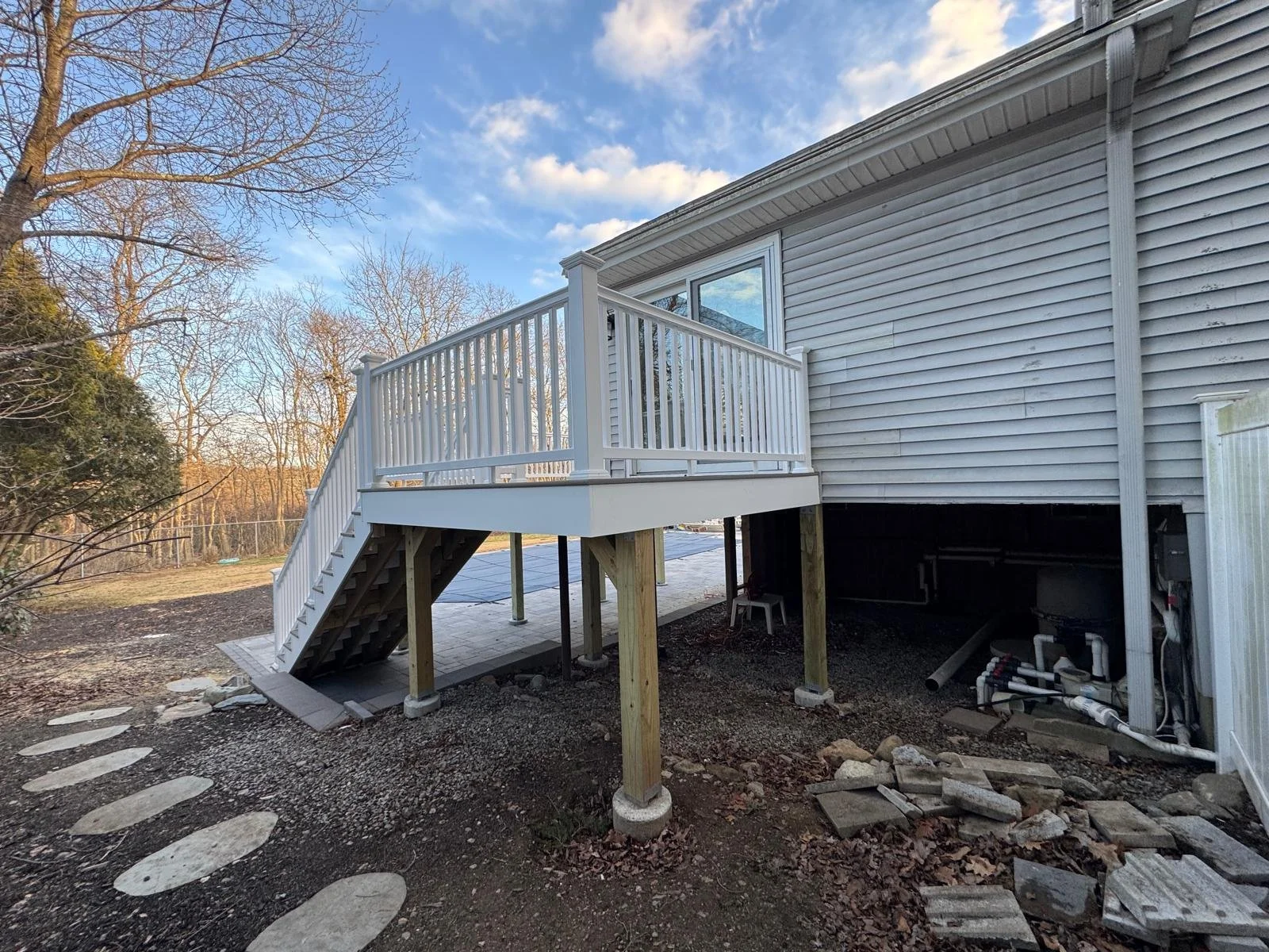 Newly built wooden deck attached to the back of a house, with stairs leading down to the yard, supported by wooden posts, in a backyard with trees and a partly cloudy sky.