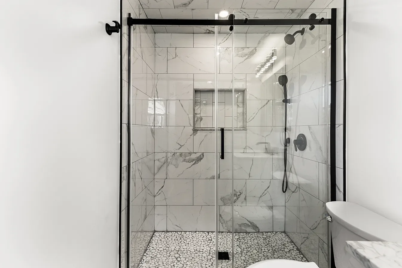 Modern walk-in shower with black fixtures, marble tile walls, glass doors, and pebble tile floor, adjacent to a white bathroom wall and toilet.