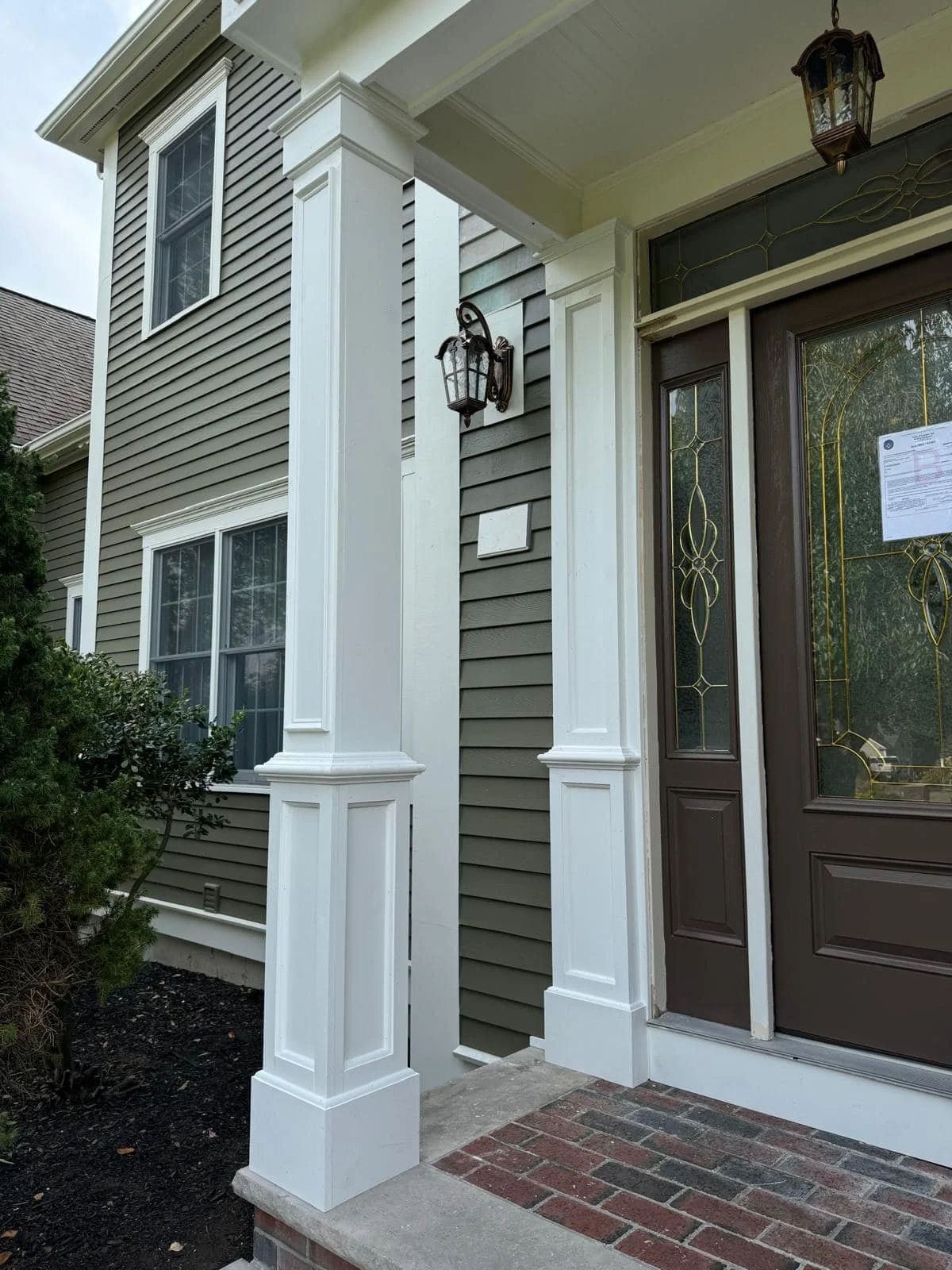Close-up view of the front porch of a house showing a decorative door with glass panes, white columns, a black lantern-style light fixture, and siding in green and white.