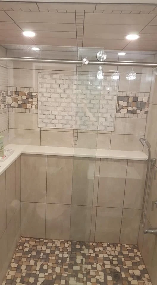 A tiled shower with clear glass enclosure, featuring a white brick accent wall, decorative mosaic tile borders on the side walls, a small shelf on the left, and a rainfall showerhead on the right.