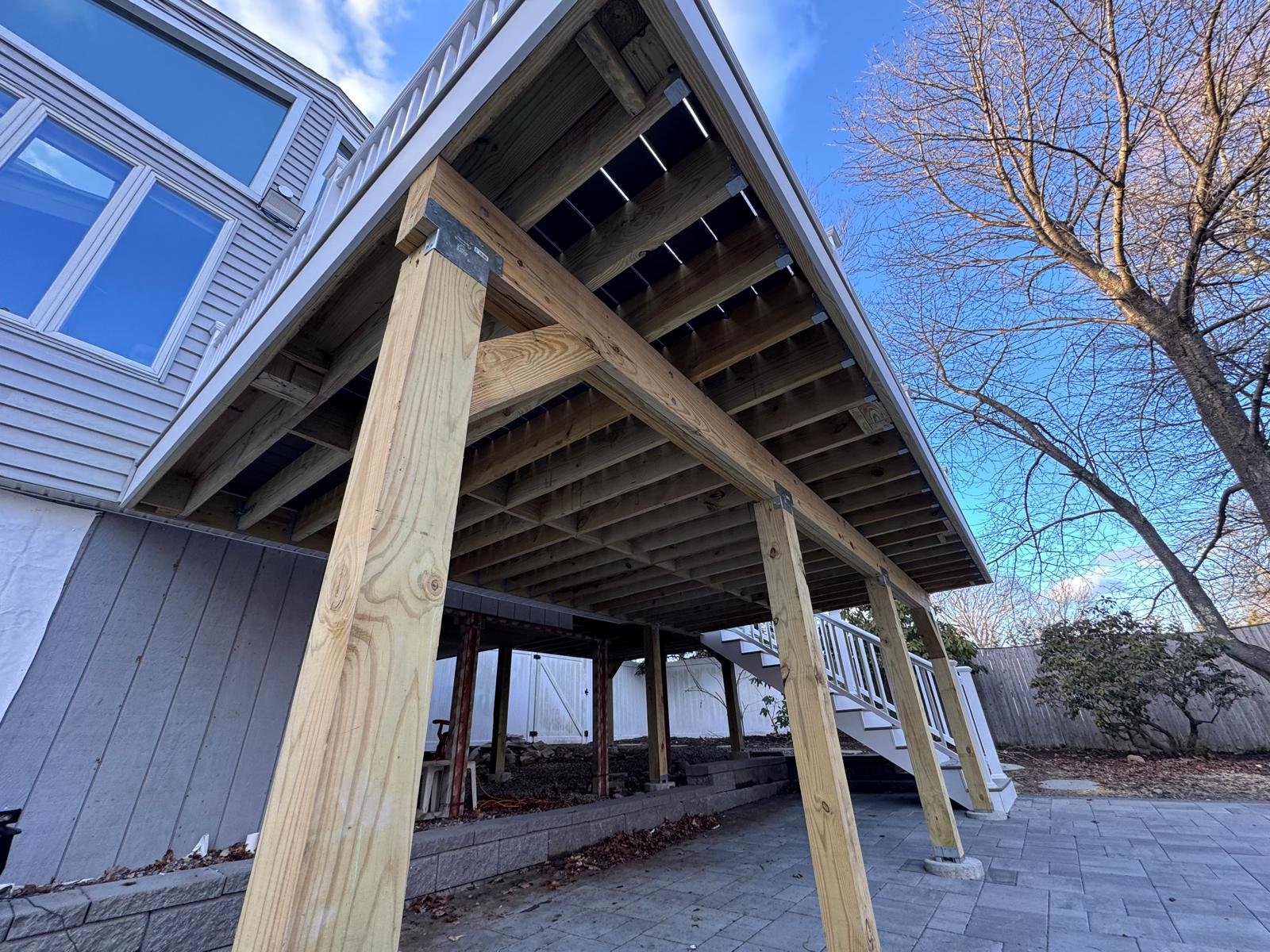 Backyard construction of a new deck with wooden support beams and stairs, neighboring a house with beige siding and blue sky with clouds.