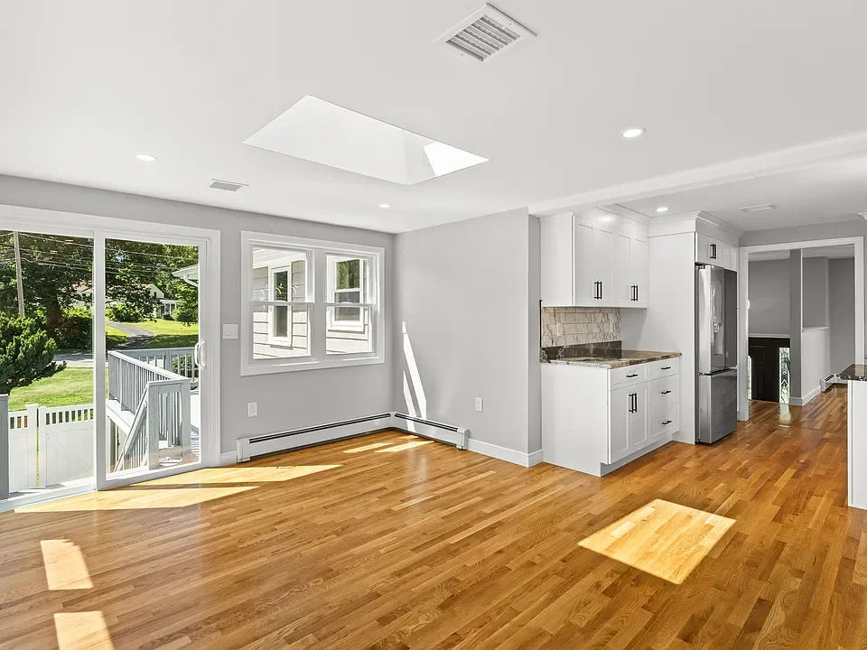 Empty kitchen and dining area with hardwood floors, white walls, large windows, sliding glass door leading outside, and a kitchen with white cabinets and stainless steel appliances.