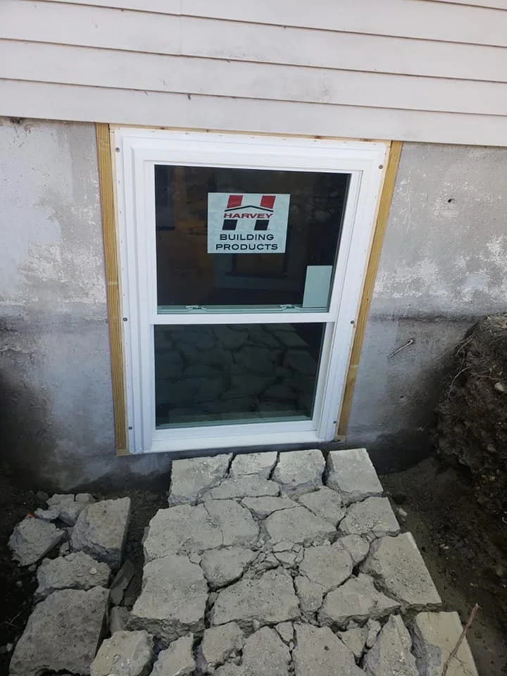 Basement window installed in a concrete foundation wall, with a new stone step in front, under construction, and partially finished siding above.