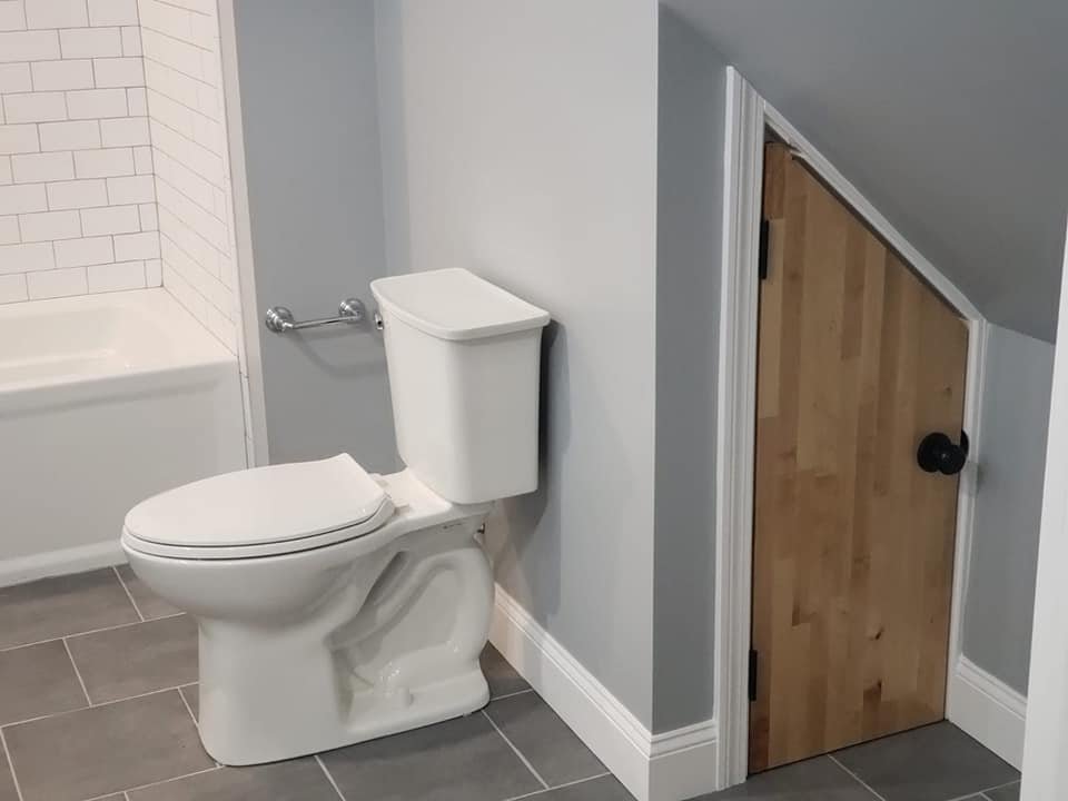 A small bathroom featuring a white toilet, a partial view of a bathtub with white subway tile, and a small wooden door with black knobs, gray tiled floor, and light gray painted walls.