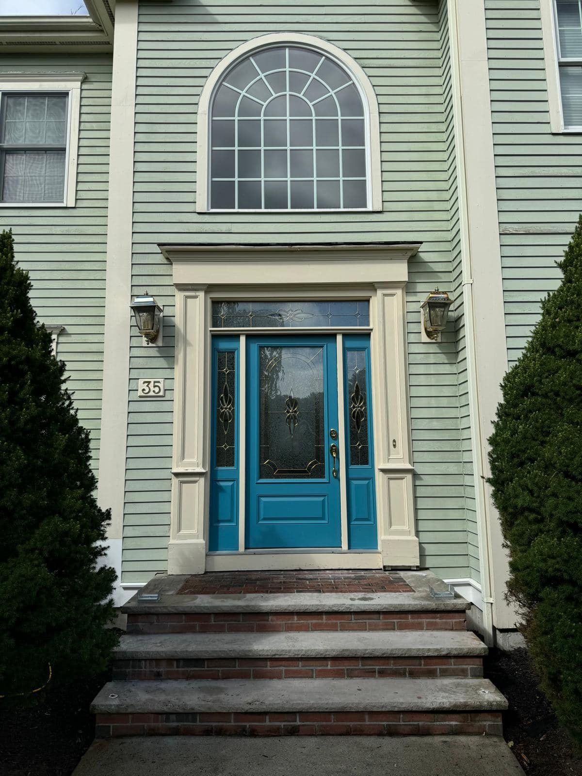Front entrance of a house with a blue door, brick steps, surrounded by green bushes, with large arched window above and exterior lights on either side.