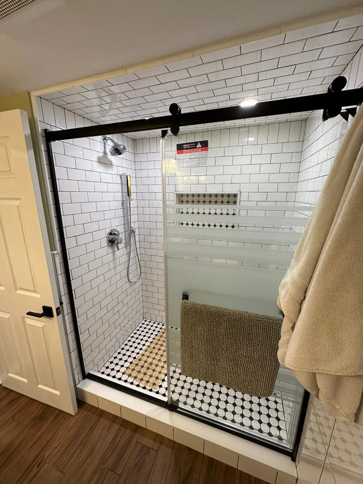A glass shower enclosure with sliding doors, white subway tile walls, black and white hexagonal floor tiles, a mounted showerhead, and a brown bath mat inside.