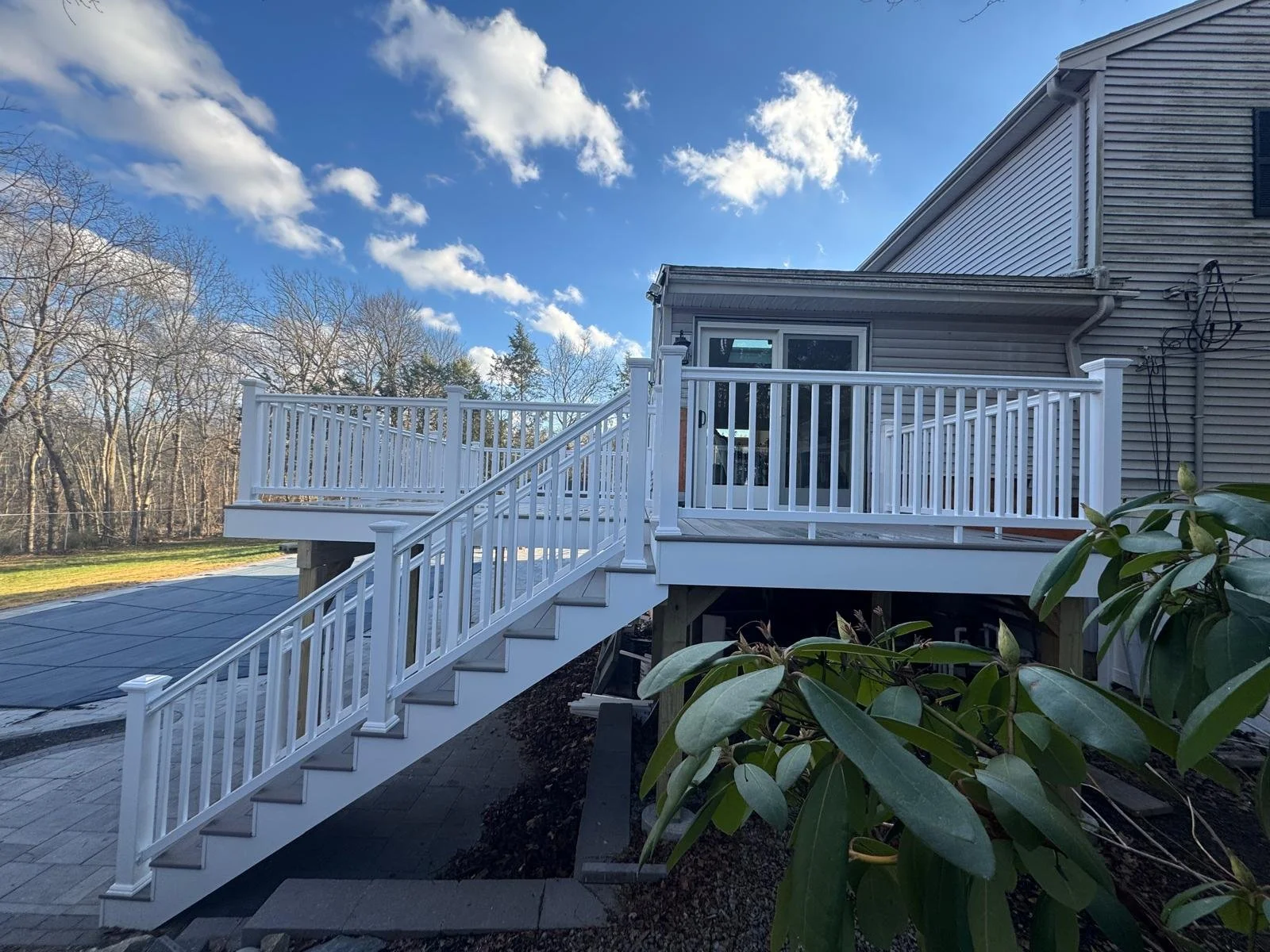 A white wooden deck with railing and stairs attached to a house with beige siding. The deck overlooks a yard with trees and a blue sky with clouds.