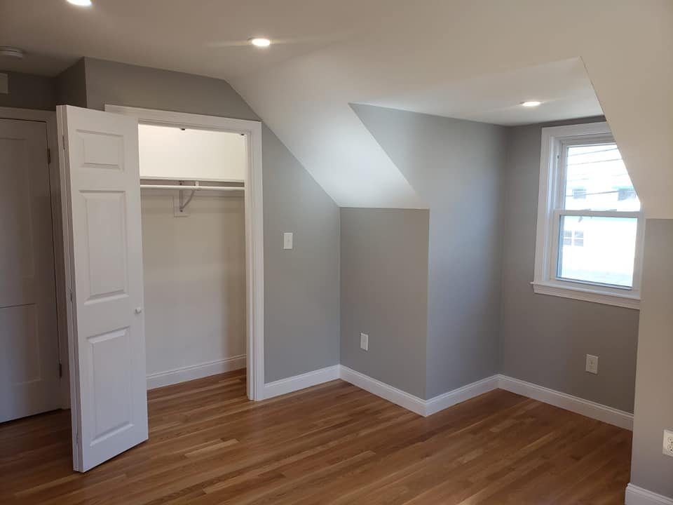 A small, empty room with gray walls, hardwood floors, and a window on the right wall. There is a closet with white double doors on the left wall, and the closet has a white interior and shelf. The room has a sloped ceiling in the corner near the wind
