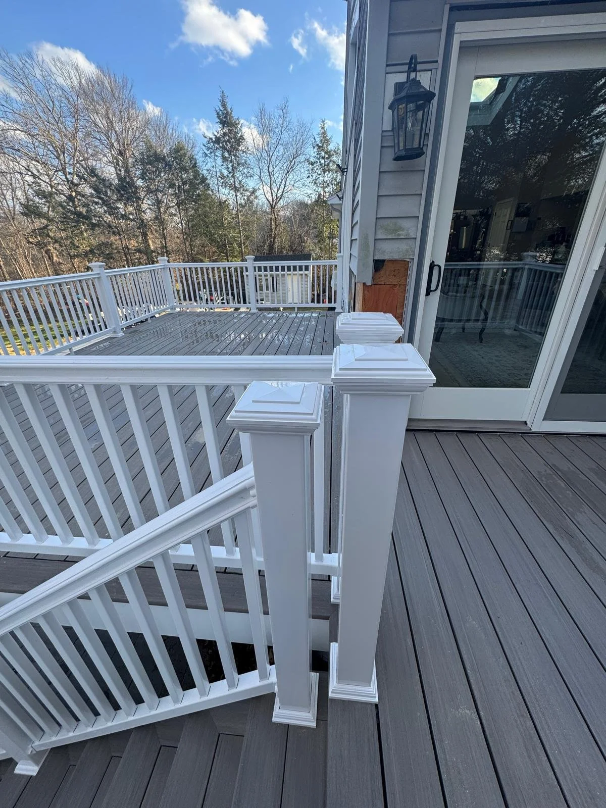 Newly built outdoor deck with gray wooden flooring, white railing, and a sliding glass door leading into the house. The deck is surrounded by a wooded area with a clear blue sky.