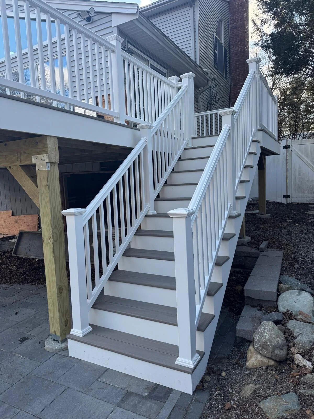 Outdoor staircase with white railing and gray steps leading to a house deck, with surrounding yard and rocks.