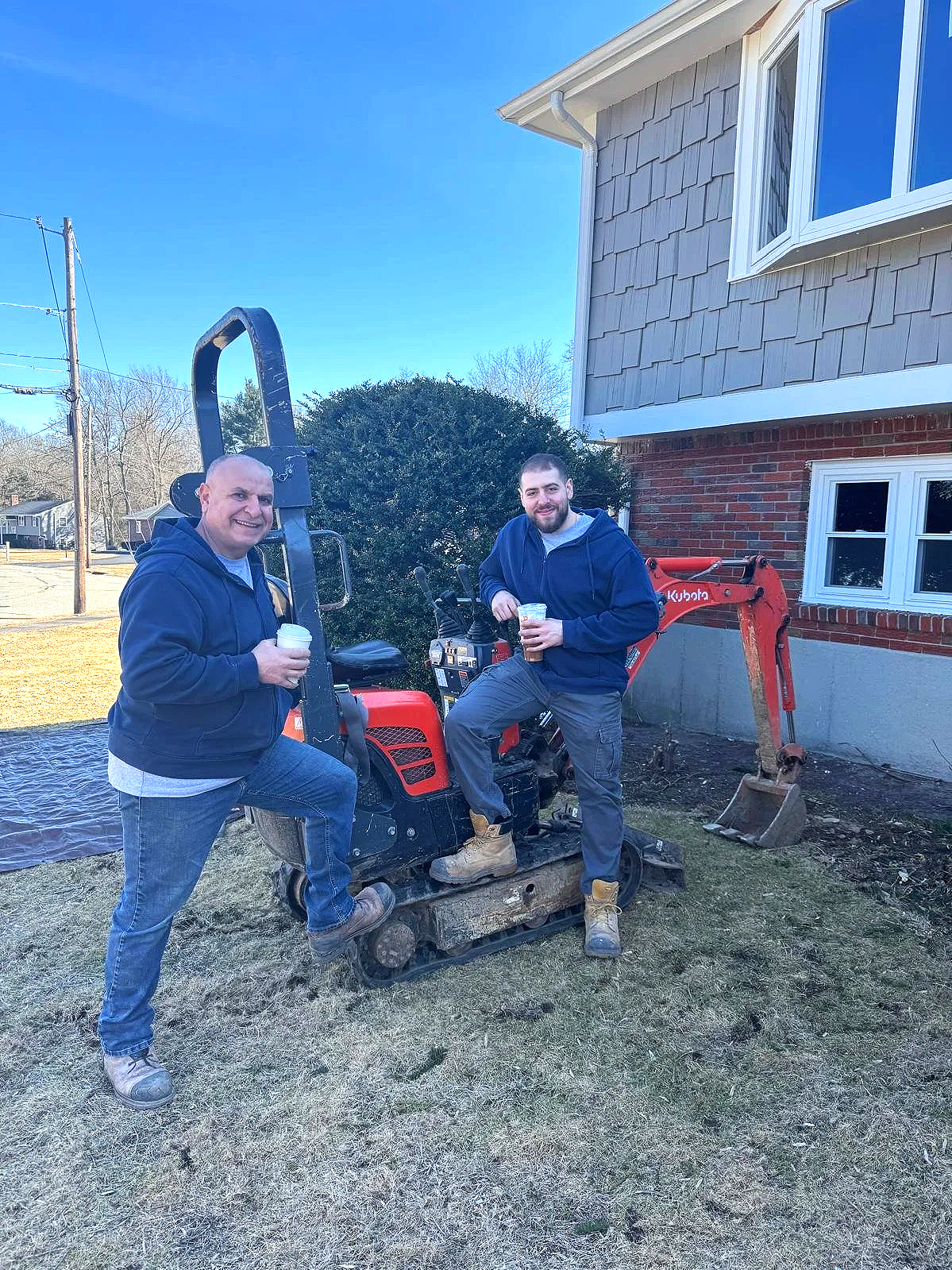 Two men smiling and holding cups of drinks, standing beside a small red excavator in a backyard with a house and a bush in the background.