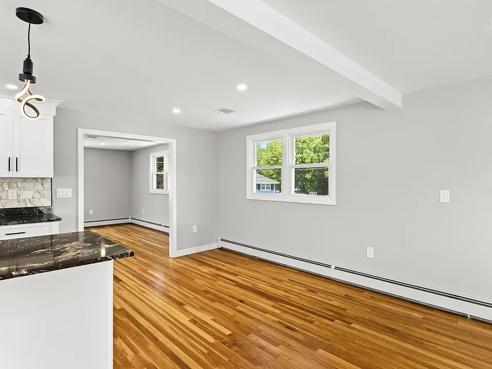 Empty living room with hardwood floors, white walls, large double-pane window, and a partial view of the kitchen.