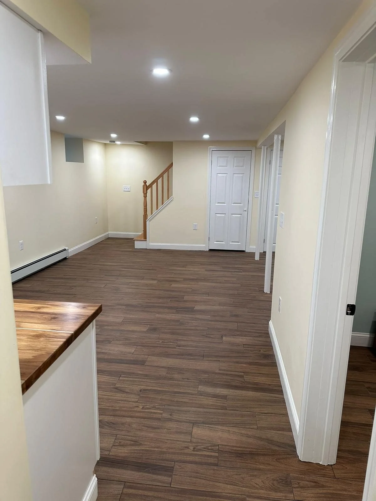 Empty living room with wood flooring, beige walls, white trim, and recessed ceiling lights. A staircase with wooden railing is visible in the background.
