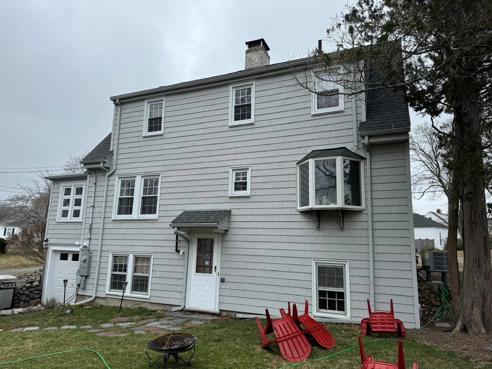 Backyard view of a multi-story house with grey siding, multiple windows, and a fire pit in the yard. There are red plastic chairs stacked upside down and a couple of garden tools nearby.