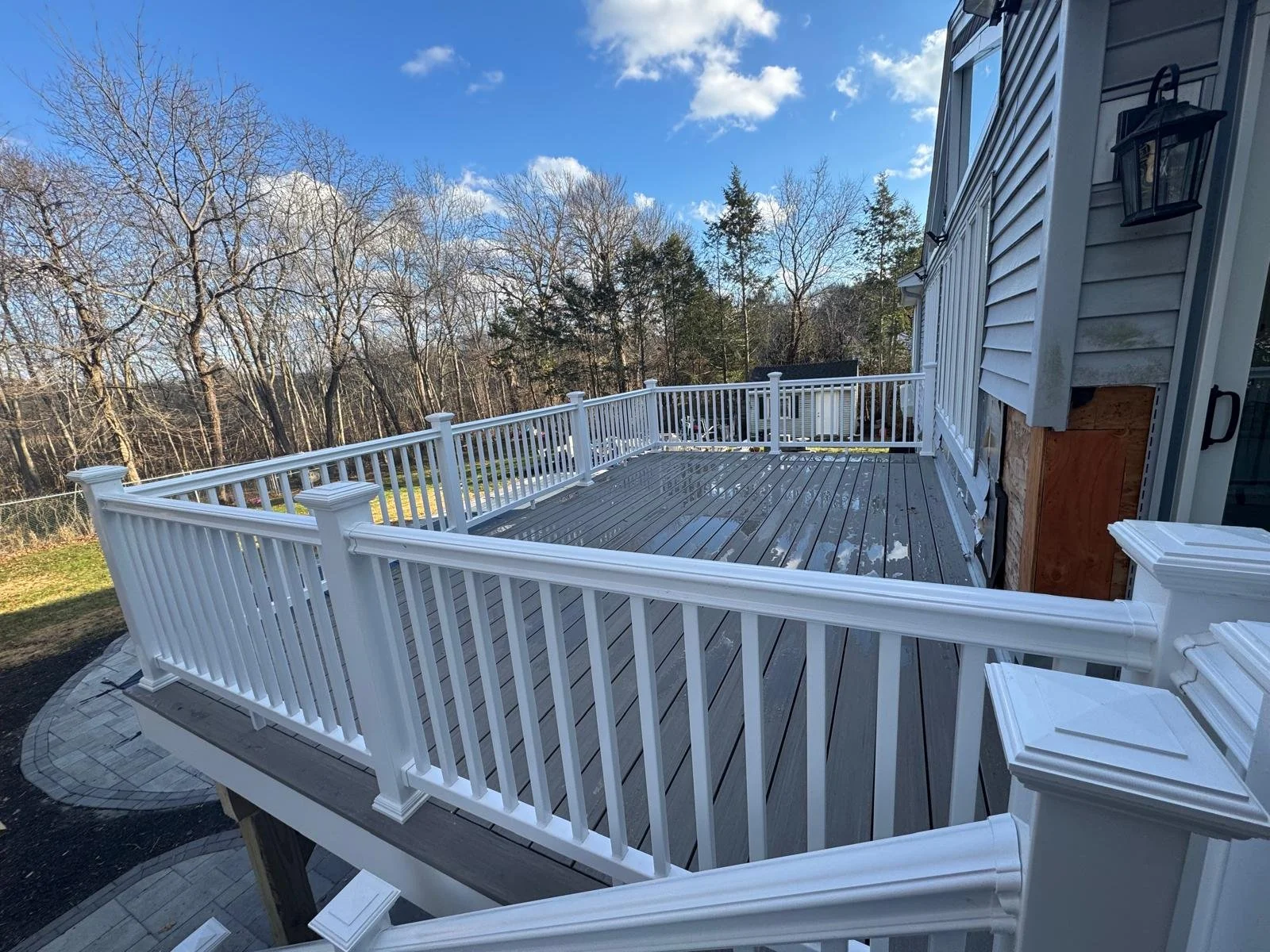View of a backyard deck with white railing, partially wet wooden planks, attached to a house with gray siding, lantern-style external light, and a wooded area with trees in the background under a partly cloudy sky.