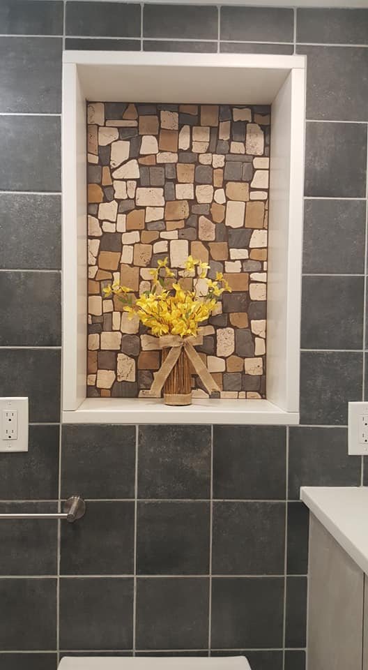 Decorative bathroom wall with a recessed niche featuring a mosaic of small stone tiles, a pitcher with yellow flowers, and gray tiled walls.