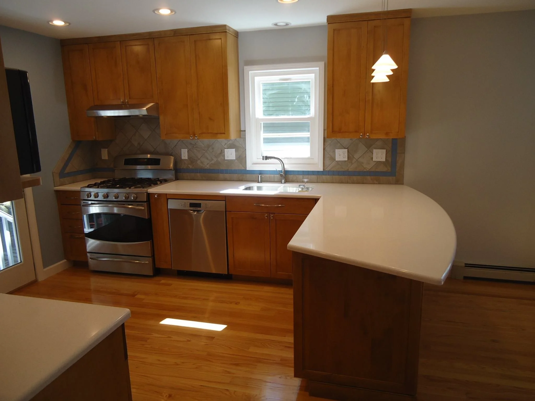 A kitchen with wooden cabinets, a stainless steel stove and dishwasher, a white countertop with a curved edge, a window above the sink, and a hanging light fixture.