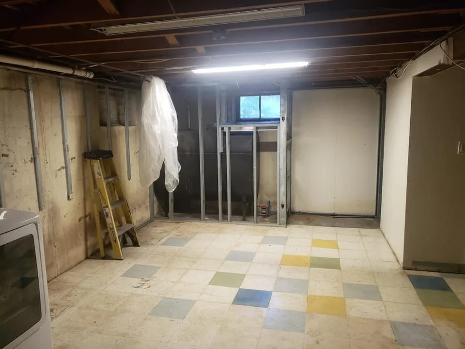 Empty basement room under renovation with exposed wall studs, ladder, plastic sheet, small window, and multicolored tile floor.