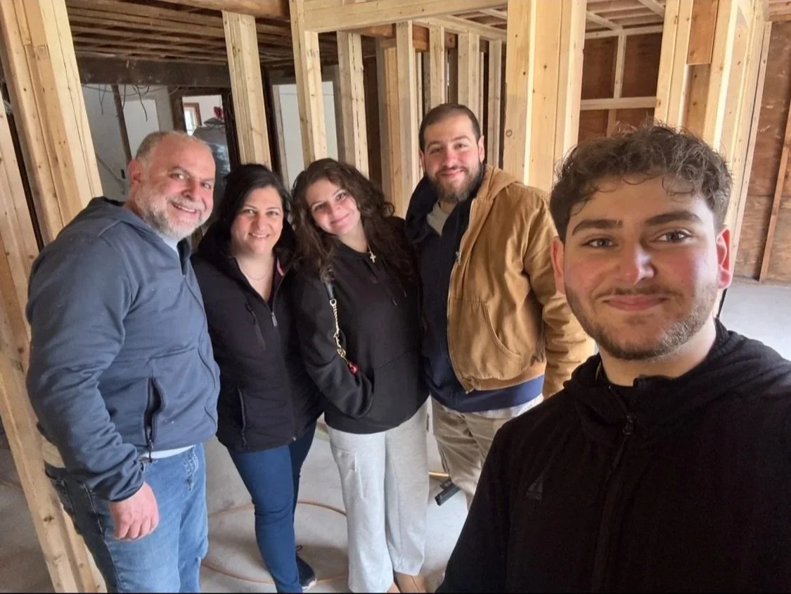 A group of five people smiling at the camera inside a partially constructed building with exposed wooden framing.