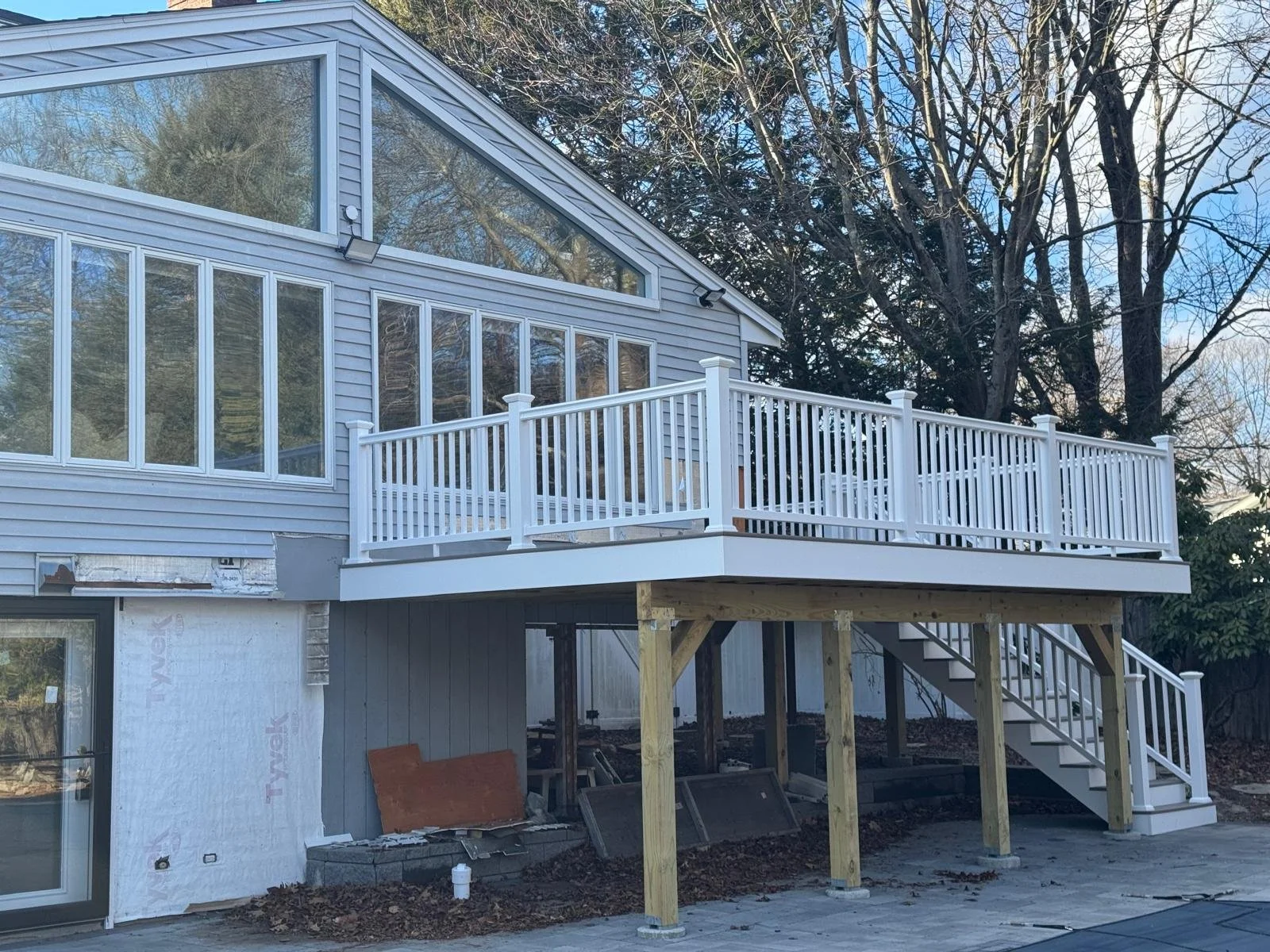 Construction of a new elevated deck attached to the back of a house, with white railing, wooden support beams, stairs, and glass-enclosed upper level. The house has light gray siding and several large windows.