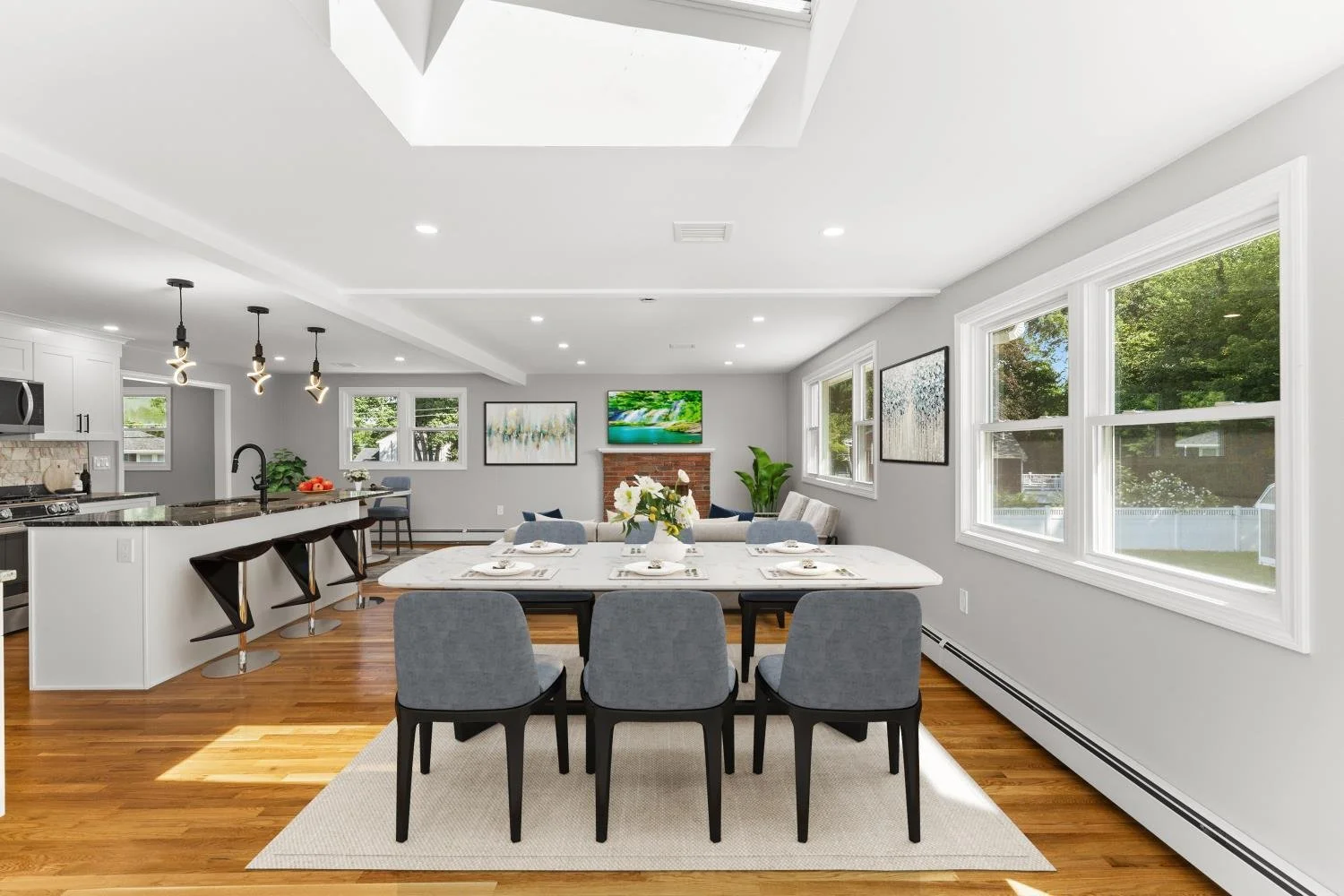 Open-concept living and dining area with large windows, hardwood floors, a white dining table with six gray chairs, a couch, a fireplace with a TV, and kitchen with barstools.