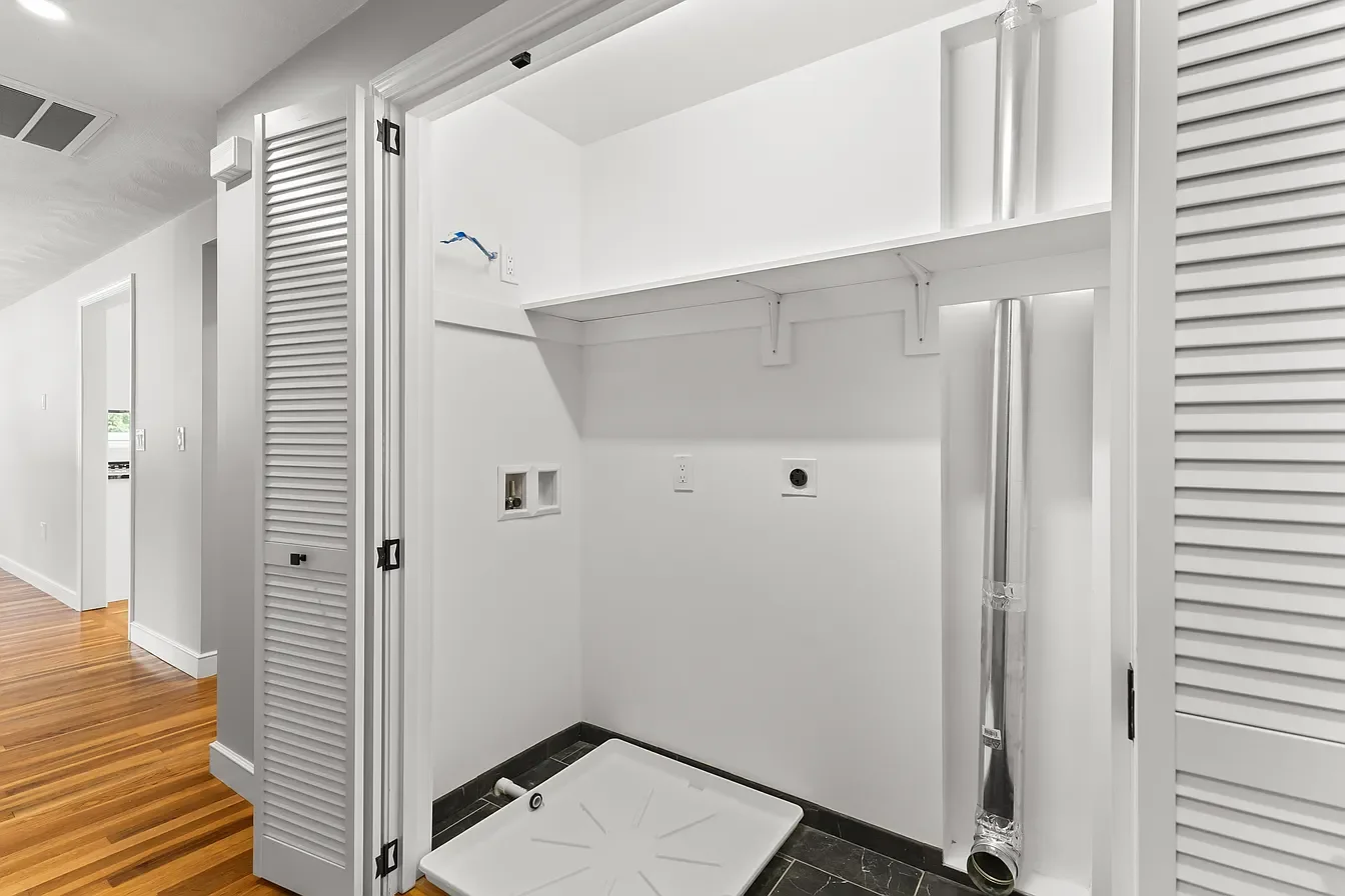 Empty laundry area with white walls, black floor tiles, and exposed vent pipe on the right, with electrical outlets and connections for washer and dryer.