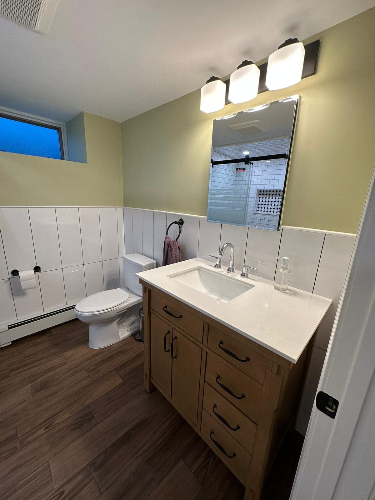 A bathroom with a vanity, mirror, toilet, and shower, featuring wood flooring, green walls, and white tiled wainscoting.