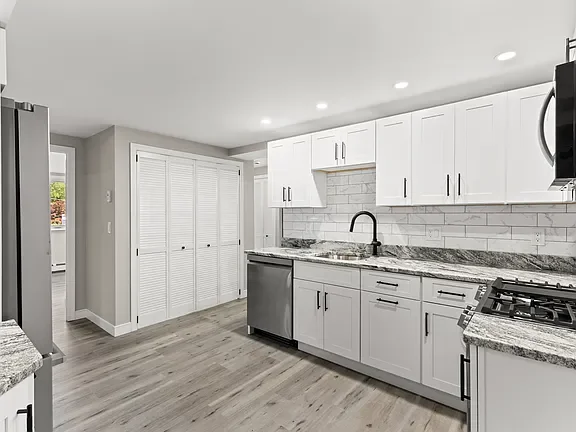 Modern kitchen with white cabinets, gray countertops, and stainless steel appliances, including a dishwasher and microwave. There is a double sink and a tiled backsplash.