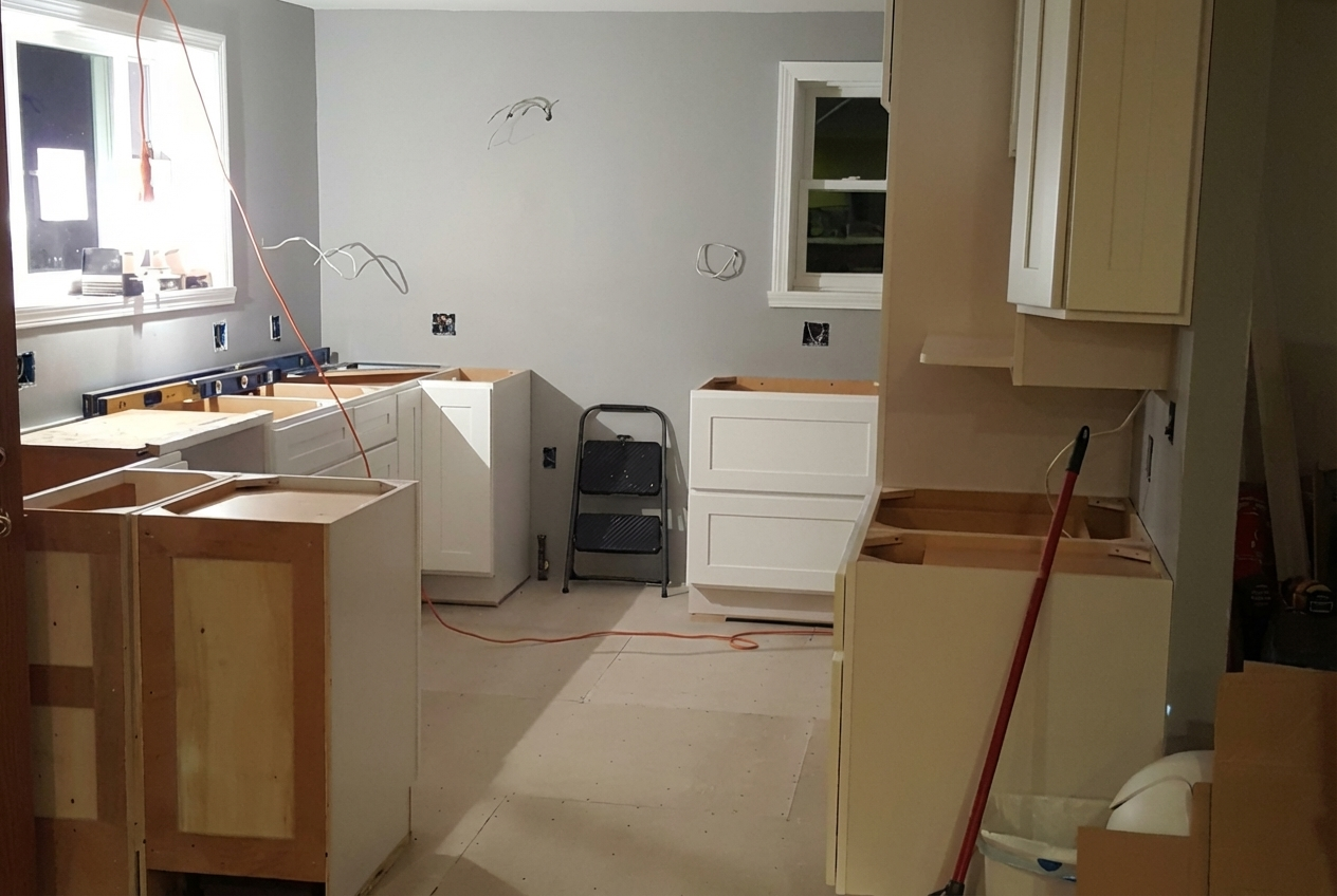 Kitchen undergoing renovation with partially installed white cabinets, a small step ladder, and construction tools, with electrical wires and outlet boxes visible.