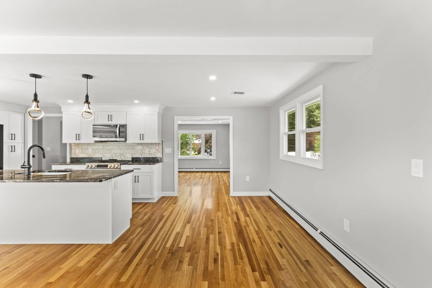 Empty open-concept kitchen and living space with white cabinets, wooden floors, and large windows