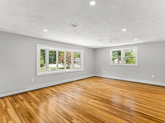 Empty living room with hardwood floors, white walls, multiple windows, and ceiling lights.