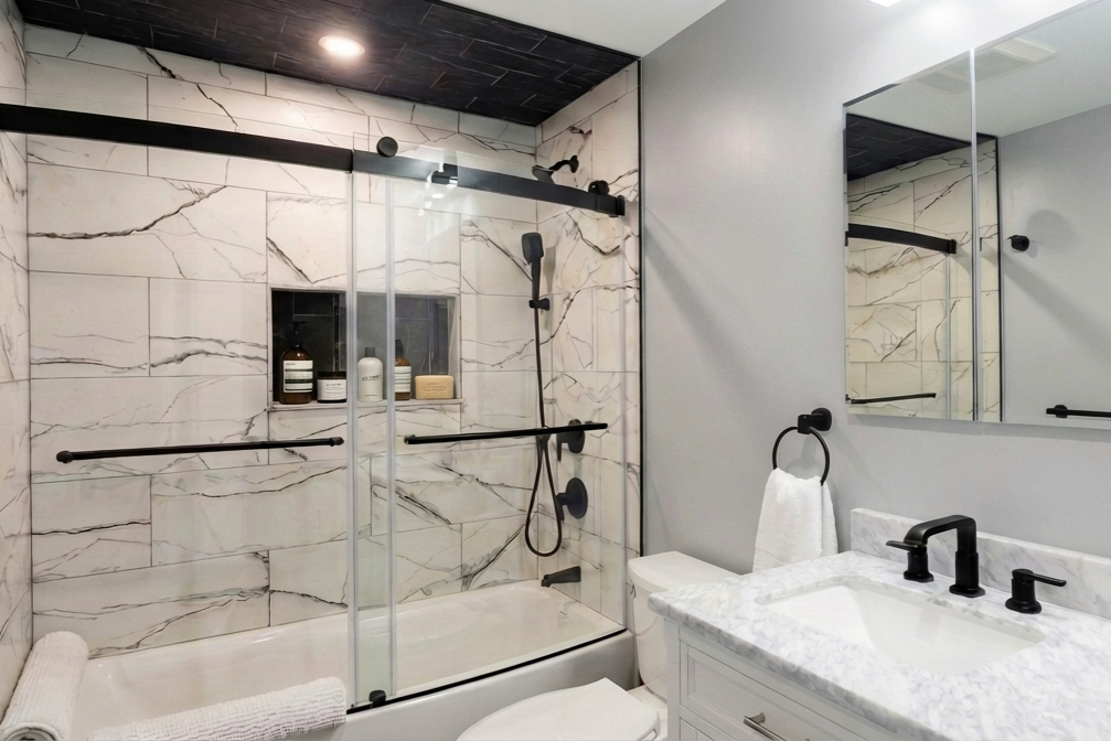 Modern bathroom with a marble shower stall, sliding glass door, and gray walls. The shower has built-in shelves with toiletries, dark fixtures, and a handheld showerhead. There is a white vanity with a marble top, black fixtures, and a large mirror.