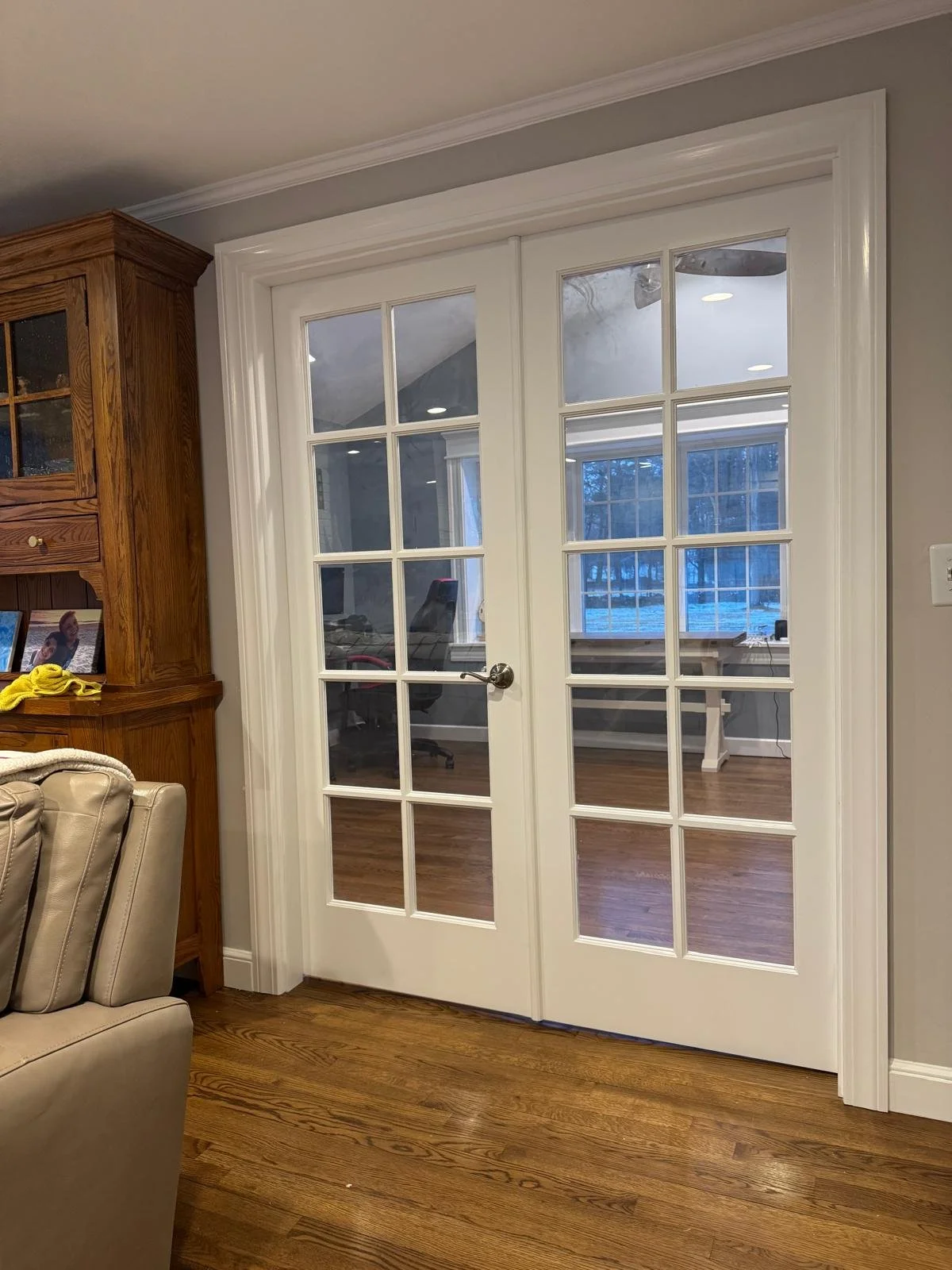 White double glass-paneled French doors with a brass handle, leading to a room with hardwood floors and large windows, in a home interior.